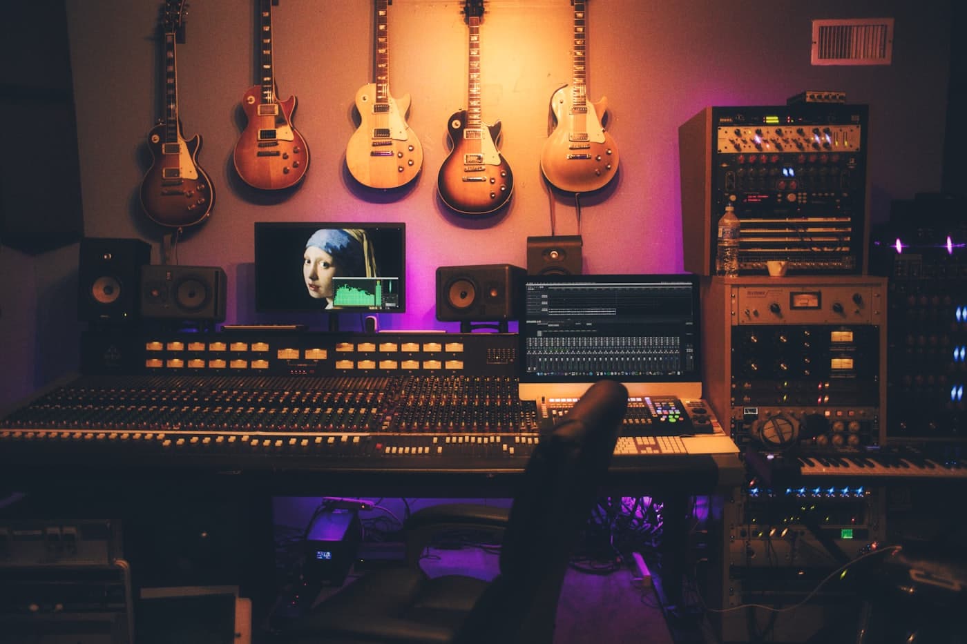 A studio control surface and mixing console with guitars racked on the wall behind it